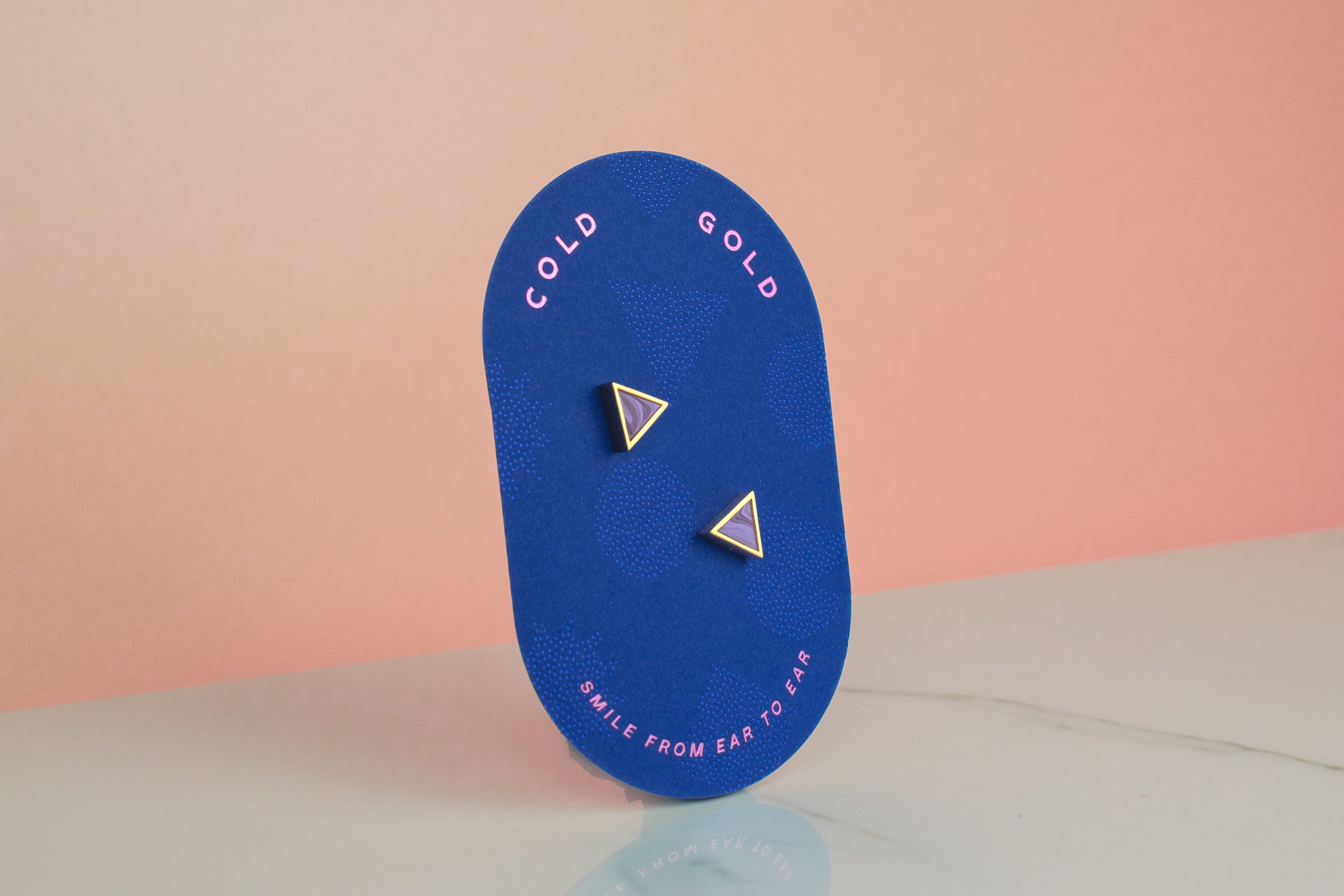 a bright blue card holding and showing a pair of gold and lavender geometric triangle earrings with 14k gold plated posts