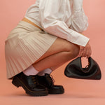 model wearing pleated skirt and chunk loafers posing with black leather crescent mini slouchy handbag