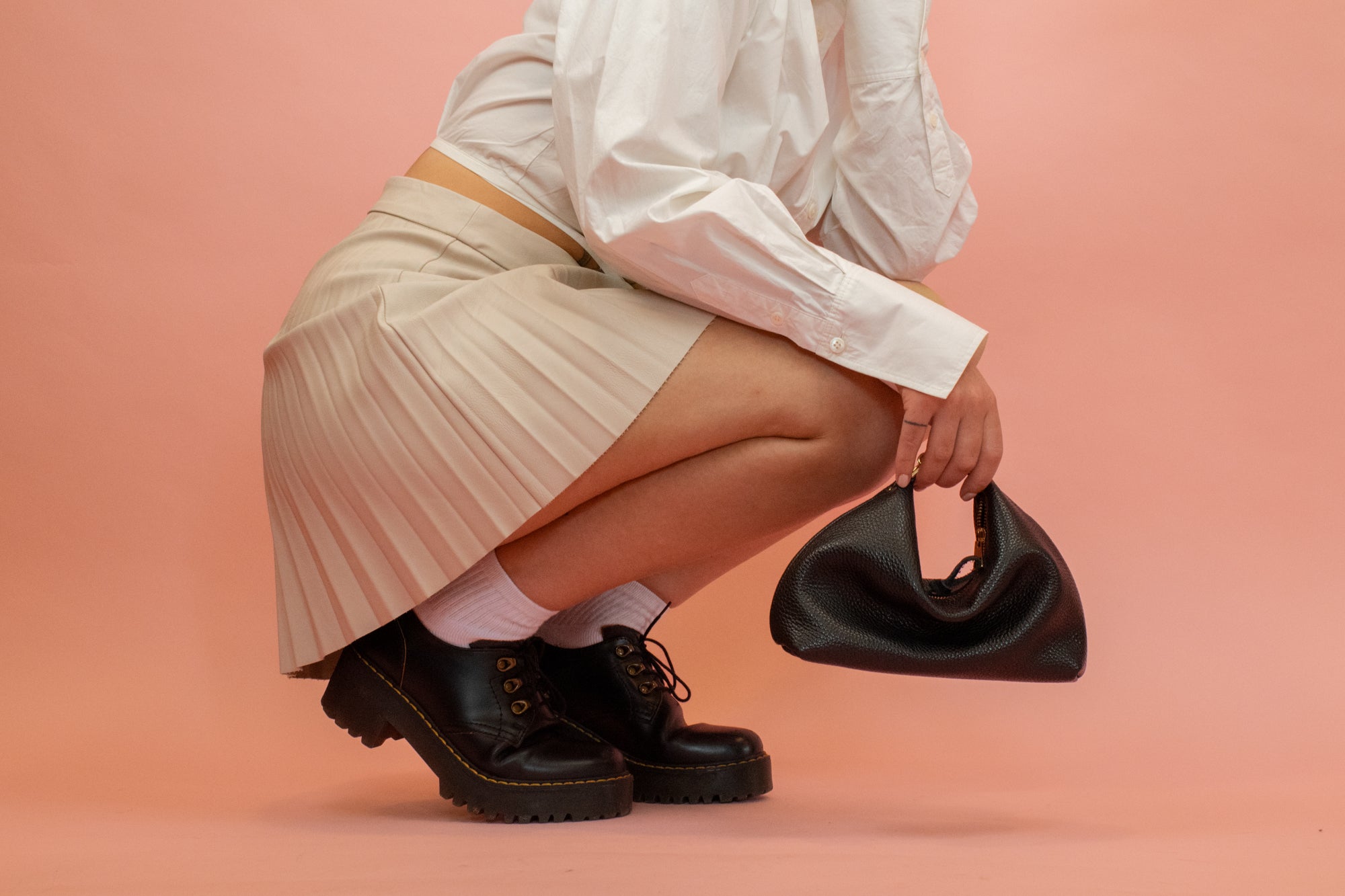 model wearing pleated skirt and chunk loafers posing with black leather crescent mini slouchy handbag