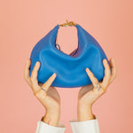 hands reaching towards the sky holding up primary blue leather mini crescent bag with relaxed silhouette and gold hardware