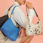 model kneeling down posing with 3 slim leather convertible handbags in blue, ivory, and cowprint