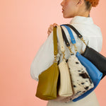 model displays 5 handmade leather shoulder bags behind her back in black, olive green, bone white, cowprint, and matisse blue