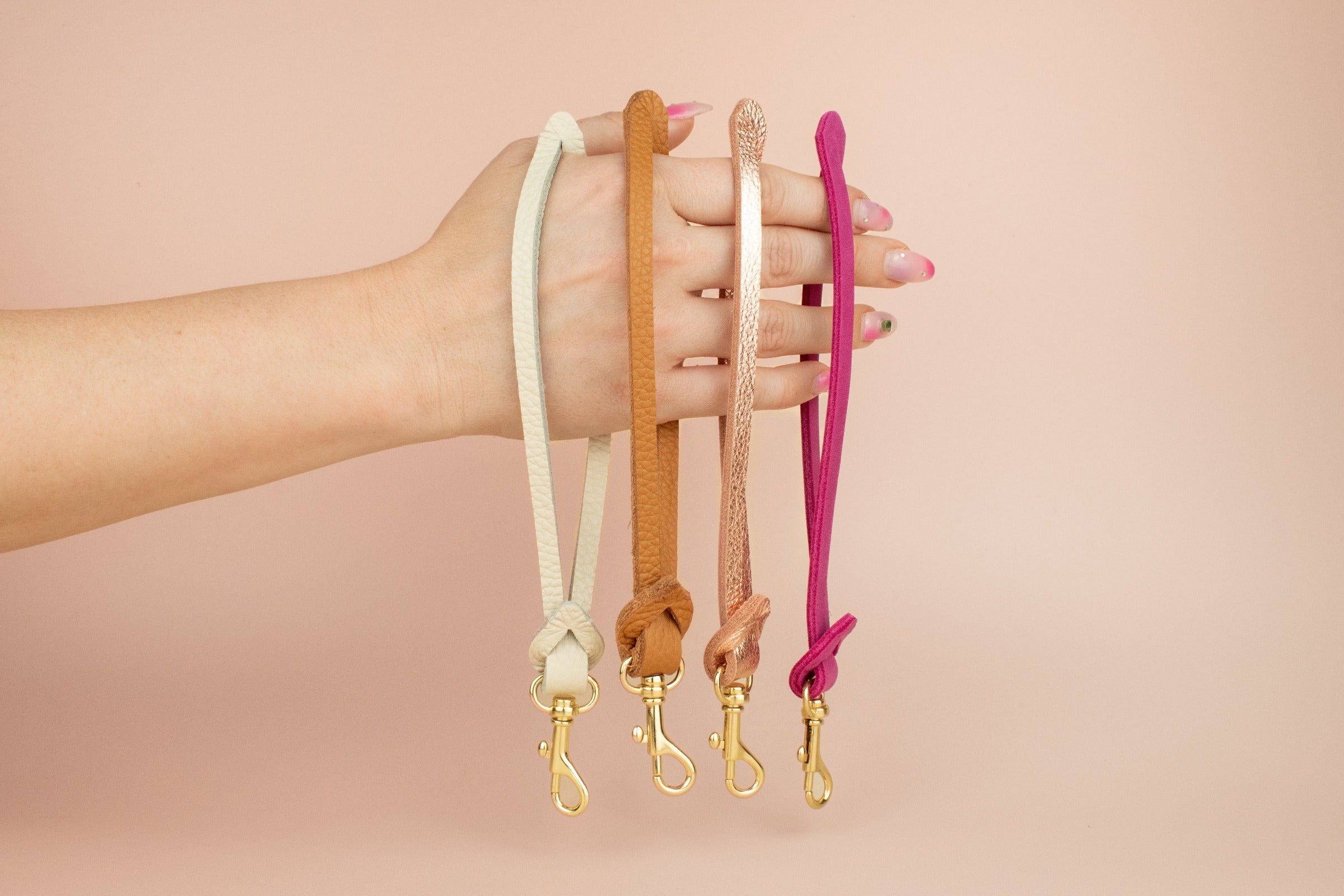 hand displaying four leather wristlet keychains with gold hardware in bone white, caramel, rose gold, and magenta pink. modern colorful purse wrist straps