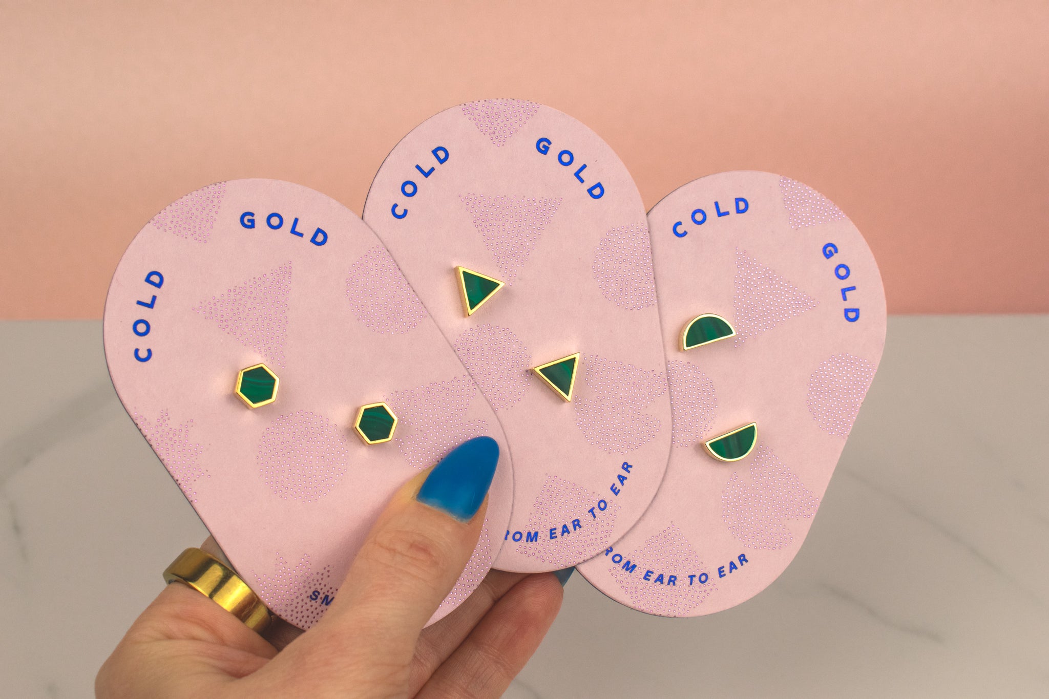 hand holding a set of three pairs of gold geometric stud earrings. One set of triangle studs, one set of gold hexagon earrings and one set of half moon stud earrings, all in emerald gemstone that's been marbled