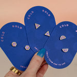 hand holding three pairs of modern and minimal pastel purple earrings in three shapes: hexagon studs, triangle studs, and half-moon studs with gold details.