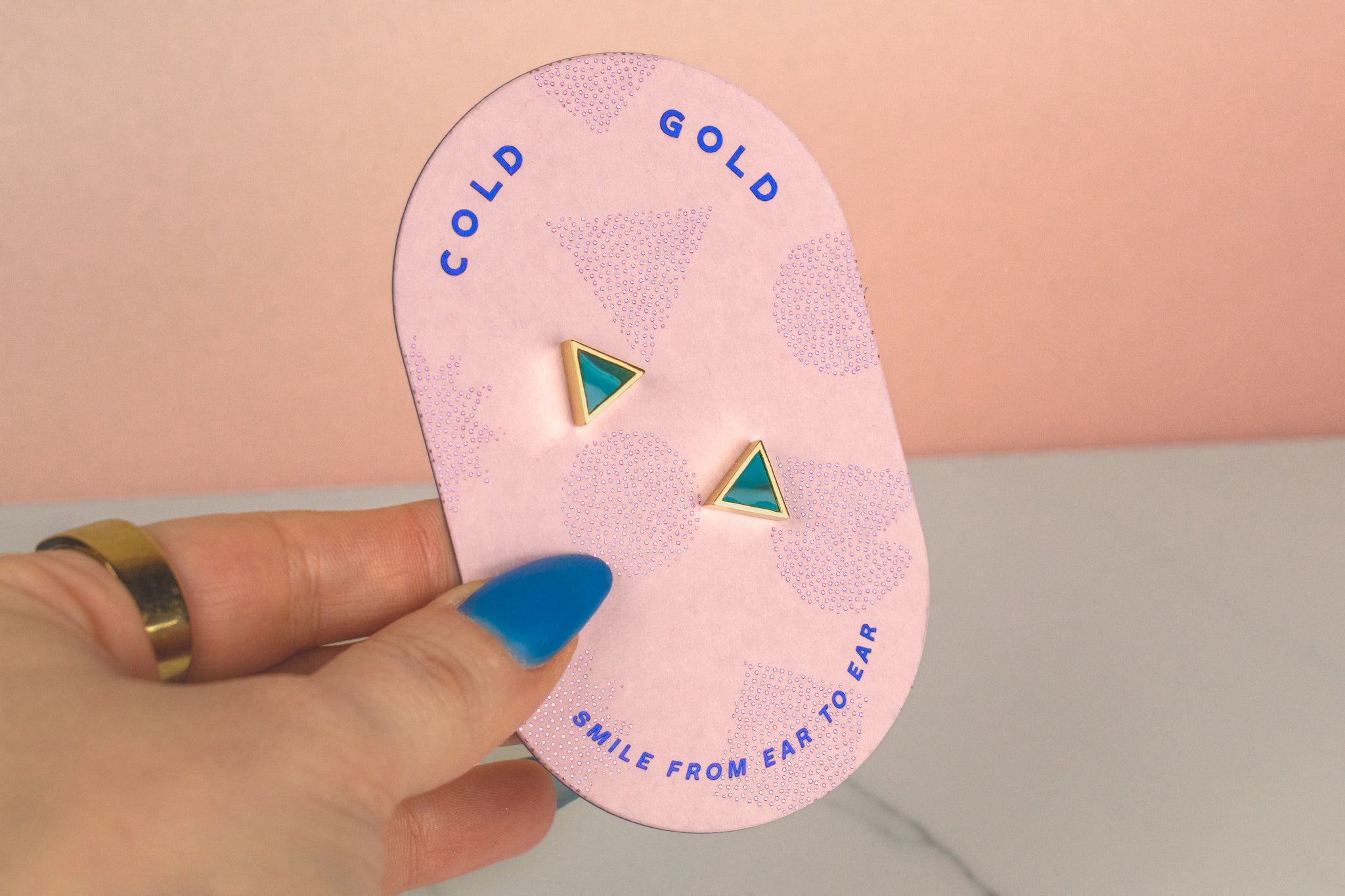 a hand holding a pill shaped pink card with a pair of marbled teal clay triangle earrings in gold