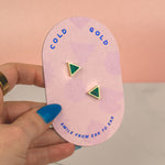 a hand holding a light pink card that shows gold branding and the cold gold logo and displaying emerald studs in a gold triangle shape that are small and dainty