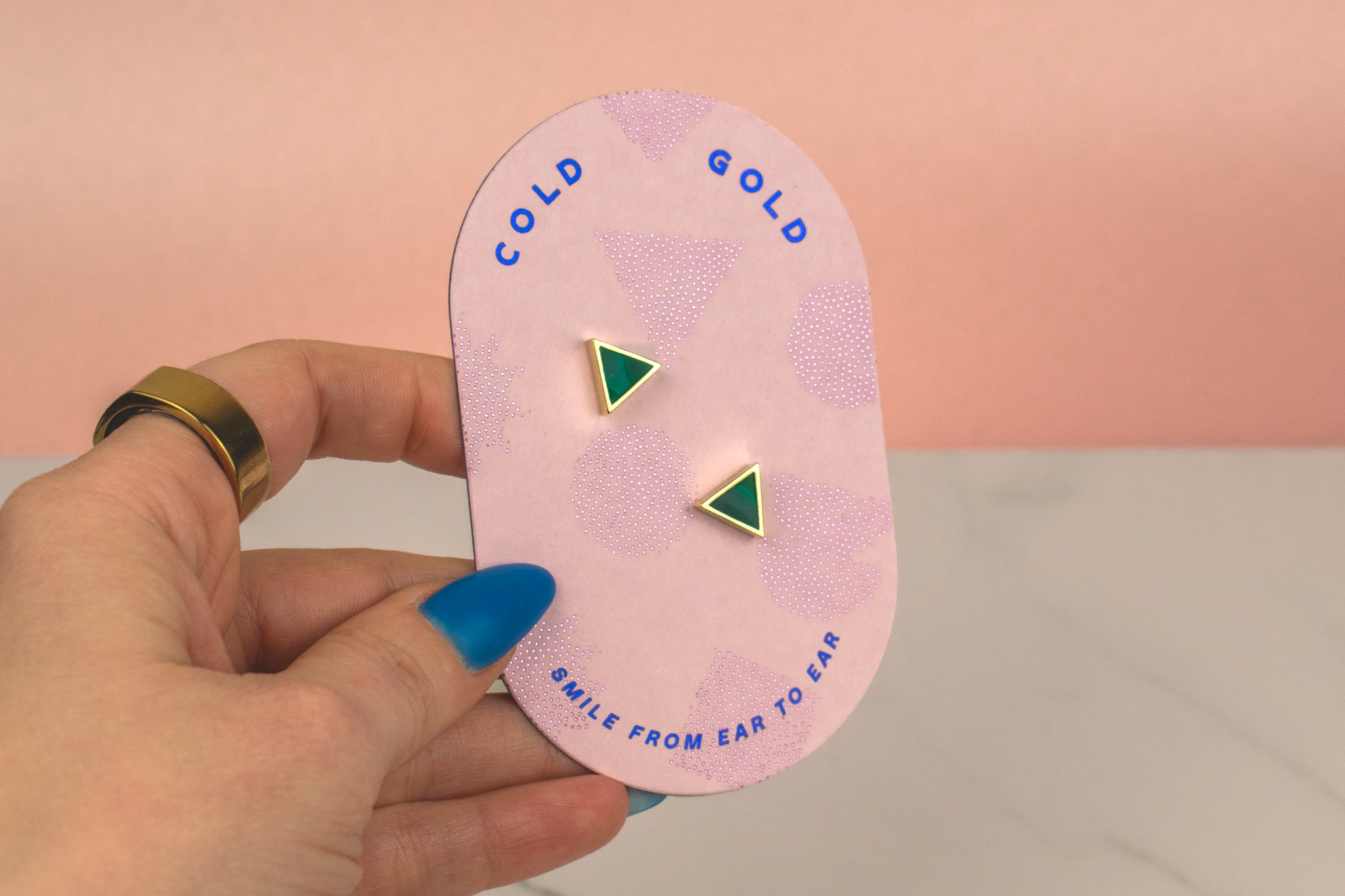 a hand holding a light pink card that shows gold branding and the cold gold logo and displaying emerald studs in a gold triangle shape that are small and dainty