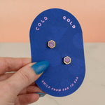 a hand holding a blue pill shaped card showing amethyst purple and gold plated hexagon geometric earring se