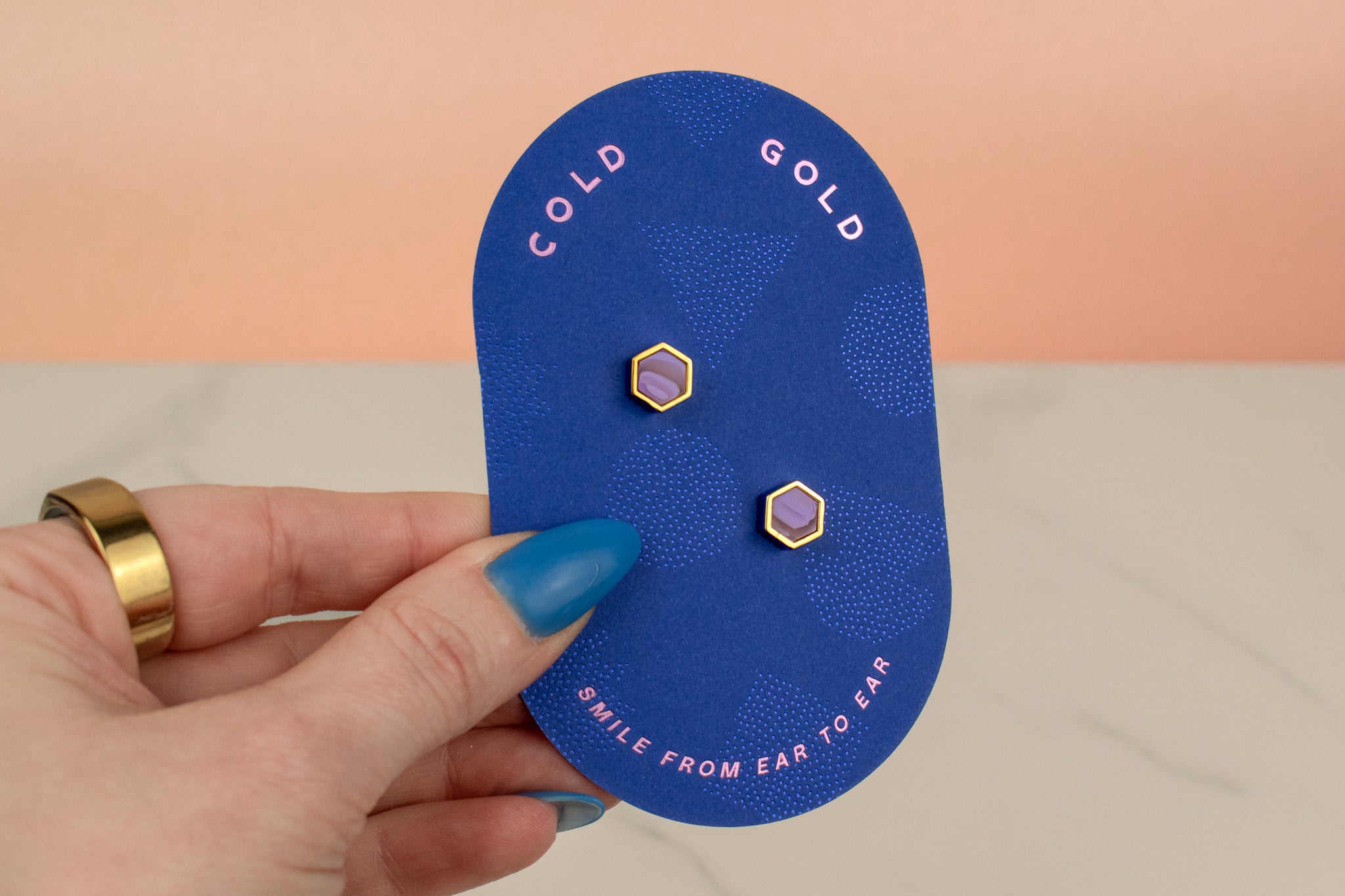 a hand holding a blue pill shaped card showing amethyst purple and gold plated hexagon geometric earring se