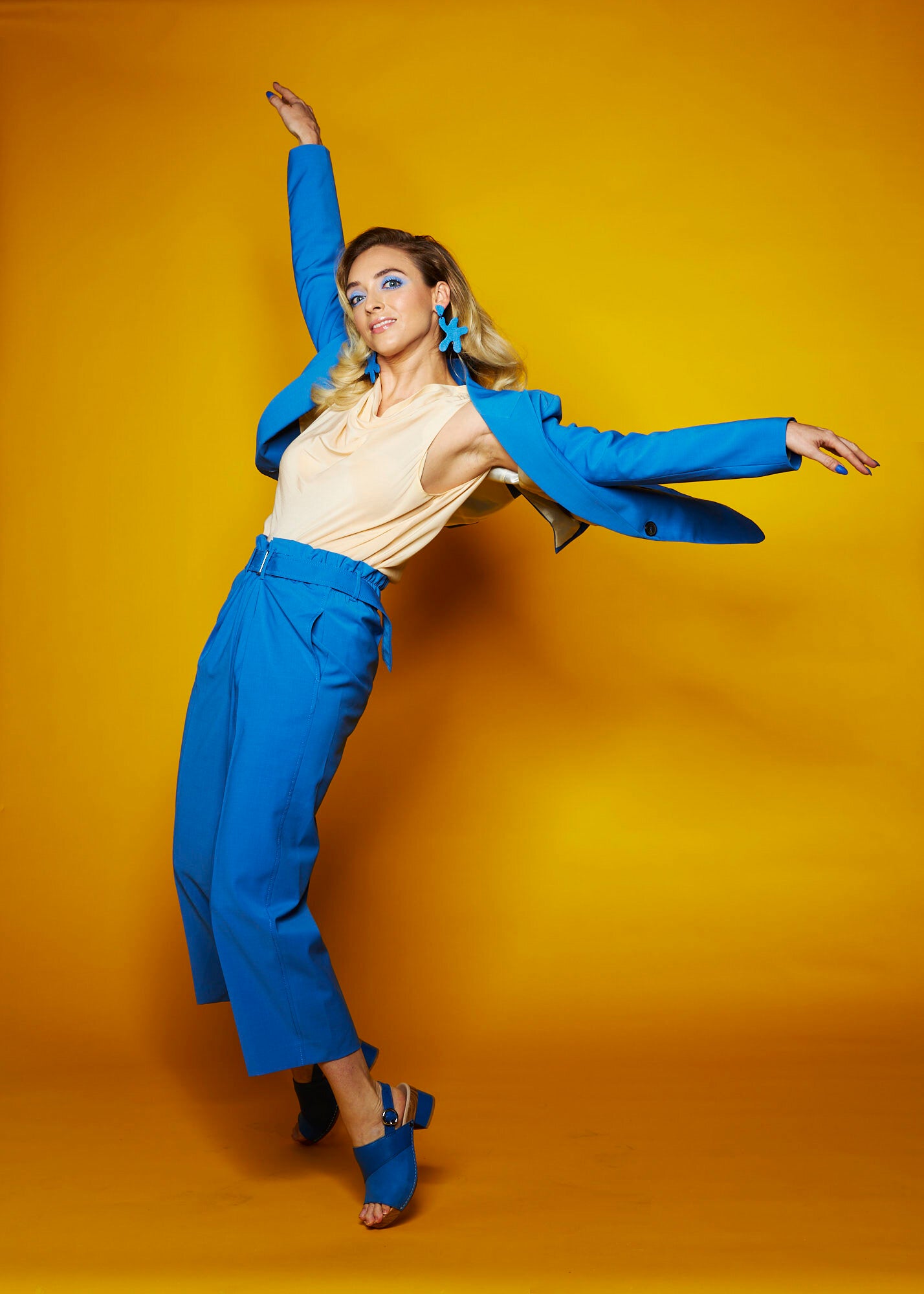 woman doing a dance pose in a blue suit wearing oversized cutout matisse earrings in blue leather.