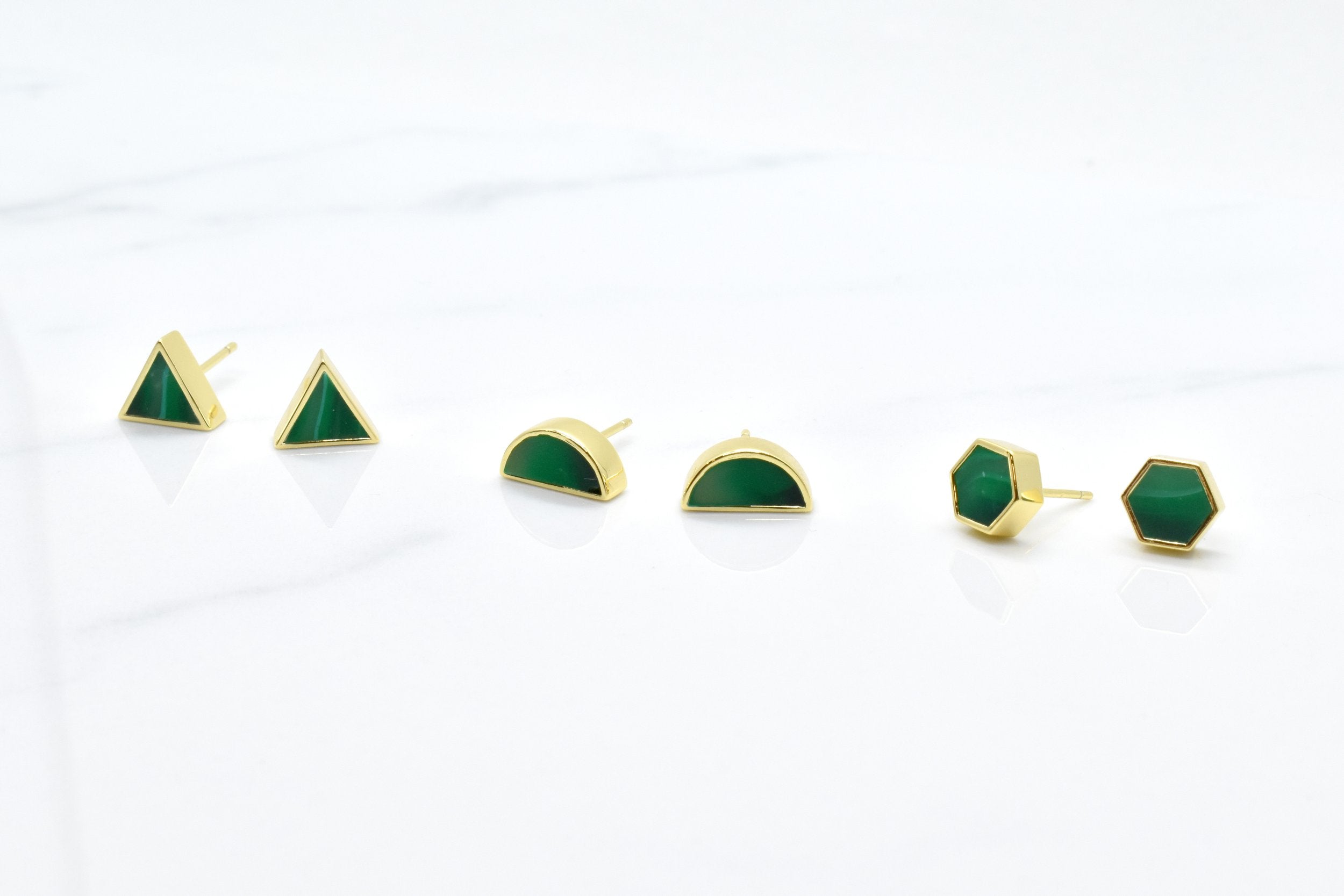 a set of three pairs of gold geometric stud earrings. One set of triangle studs, one set of gold hexagon earrings and one set of half moon stud earrings, all in emerald gemstone that's been marble