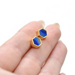 hands holding gold hexagon stud earrings with striped sapphire gemstone texture and 14k gold plated hexagon shape against a white background