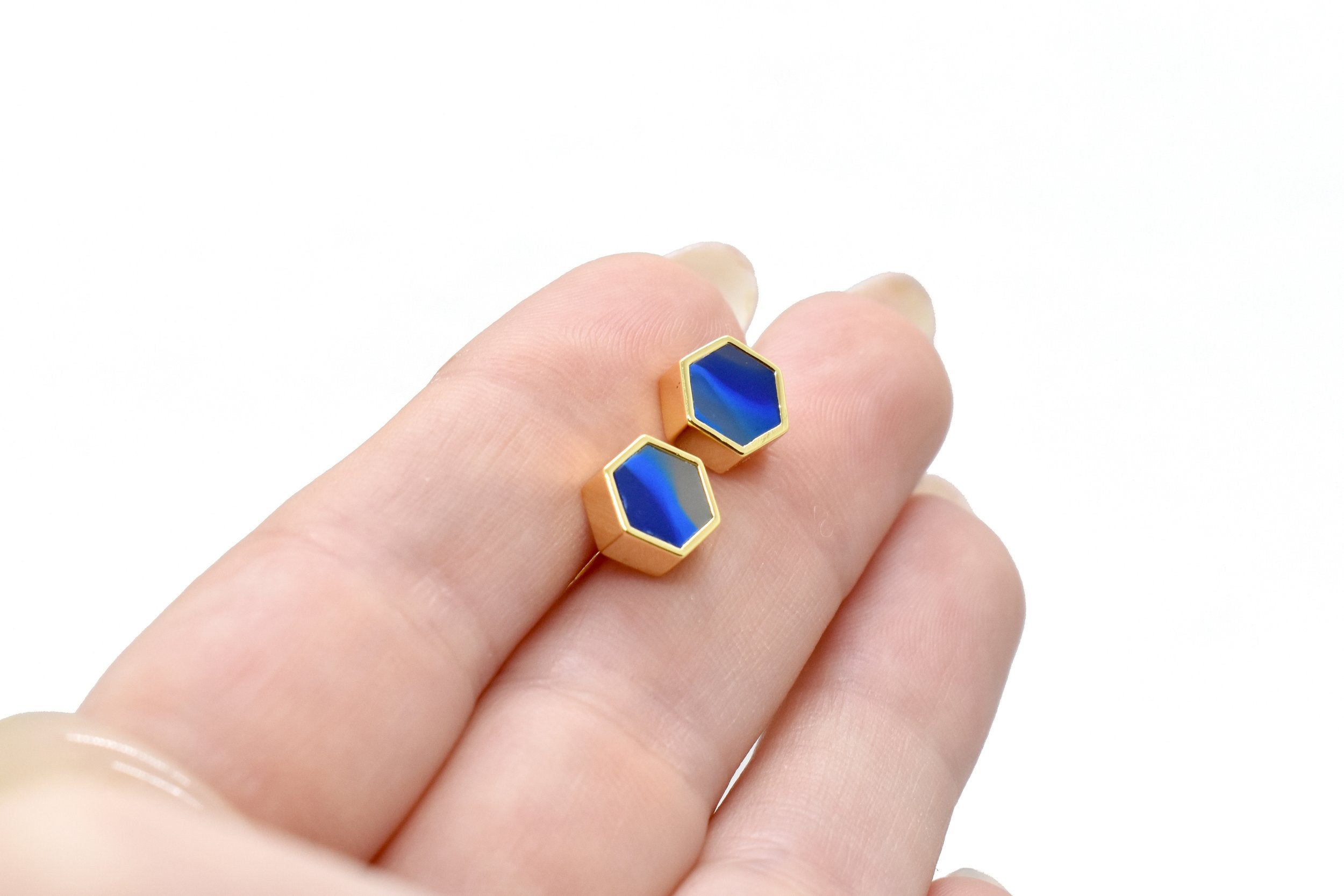 hands holding gold hexagon stud earrings with striped sapphire gemstone texture and 14k gold plated hexagon shape against a white background