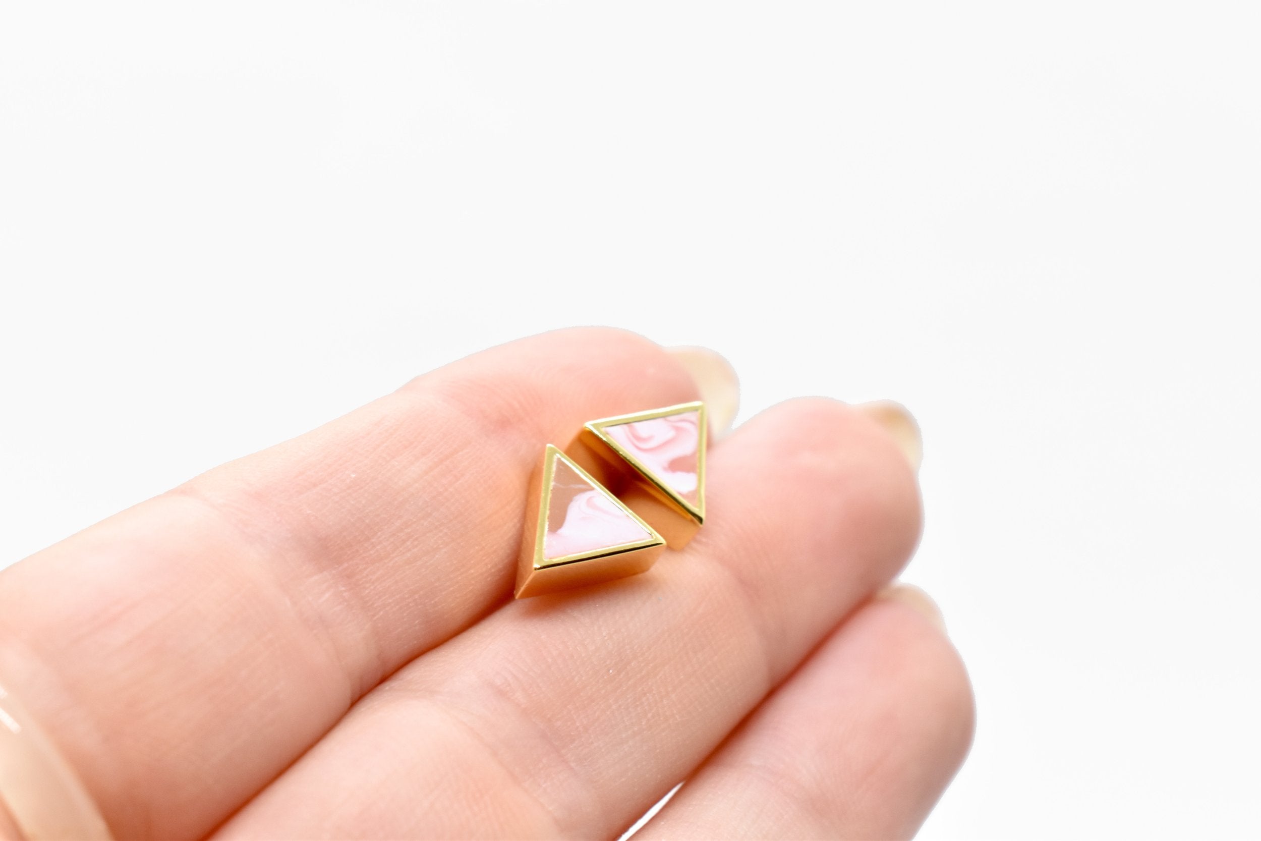 a hand holding a tiny pair of studs in pastel pink tiny triangle earrings with 24k gold plating.