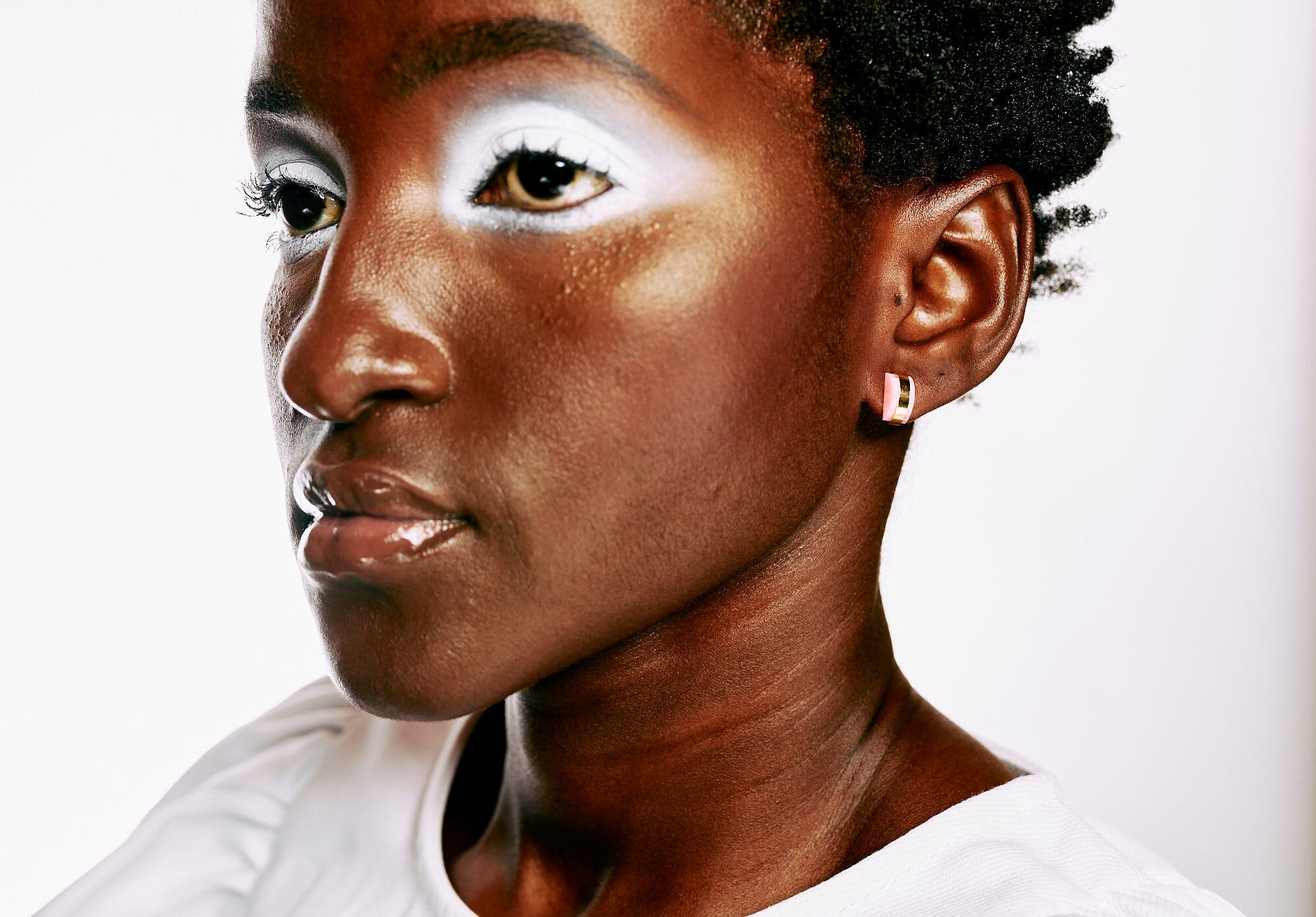 A woman with short hair and dramatic white makeup wears art-deco inspired stud earrings in semi-transparent pink clay and gold.