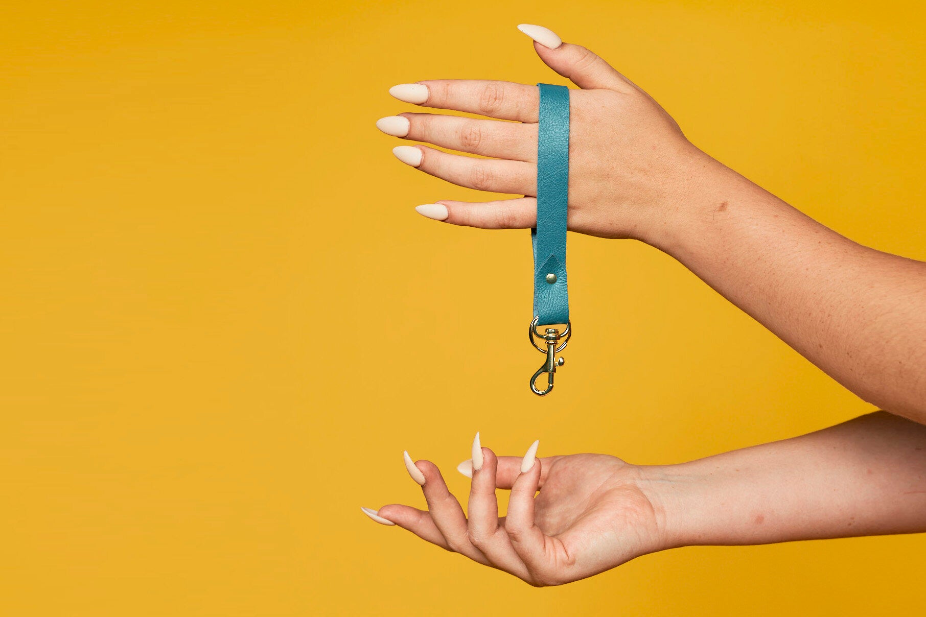 A modern key organizing wrist strap in turquoise authentic leather with gold metal detail hangs from a hand with long white fingernails.