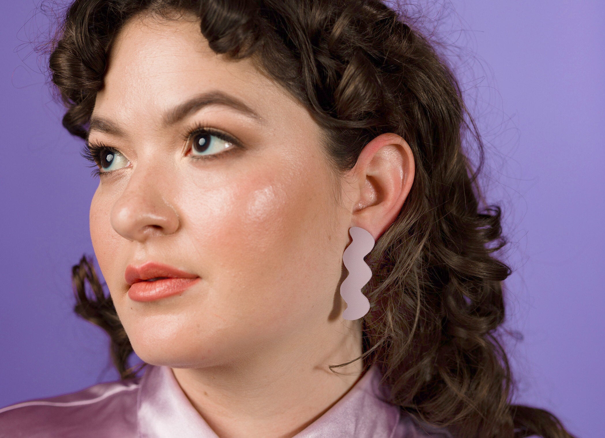 a young woman with dewy natural makeup and a shiny lavender top wears lavender leather zigzag statement earrings.