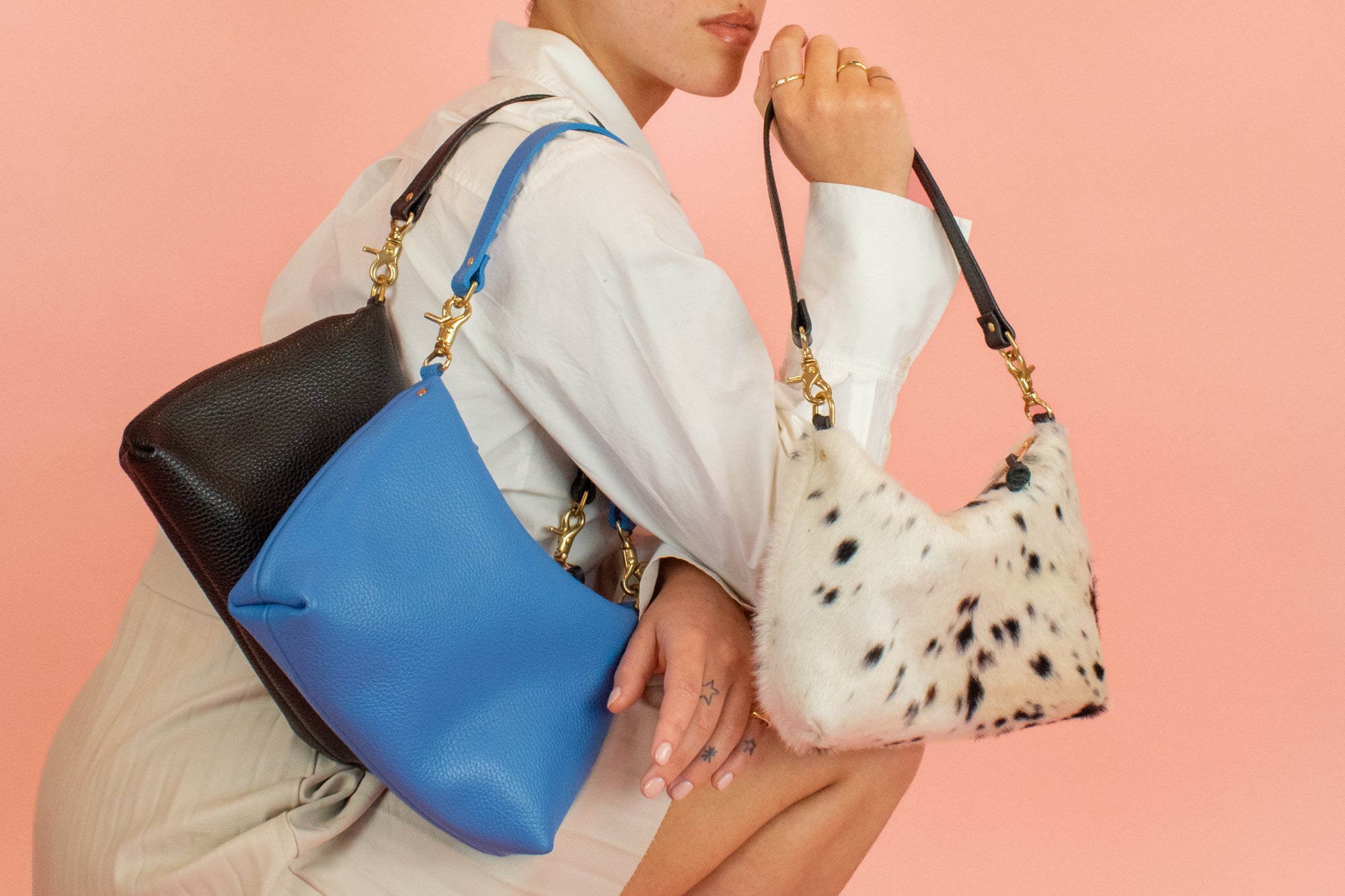 model kneeling down posing with 3 slim leather convertible handbags in blue, ivory, and cowprint