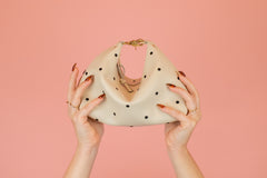Cream-colored handbag with black polka dots held by hands against a pink background