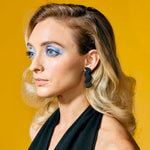 A model with blonde retro wavy hair wears the black leather Light Trace earrings.