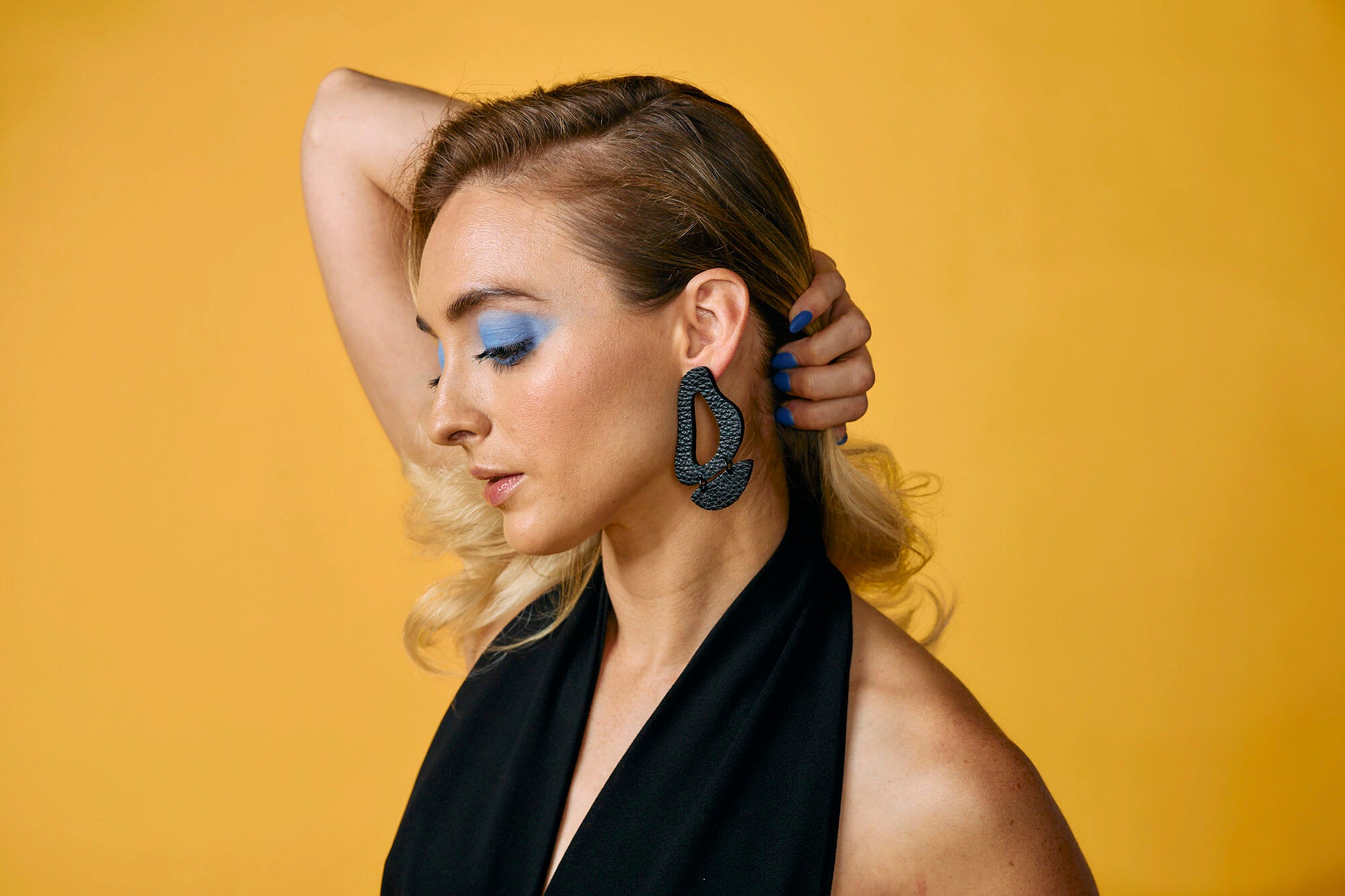 A blonde model against a yellow background poses wearing the black leather inferno statement earrings.