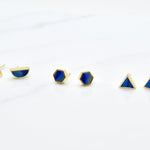 a set of three pairs of gold geometric stud earrings. One set of triangle studs, one set of gold hexagon earrings and one set of half moon stud earrings, all in sapphire gemstone that's been marble