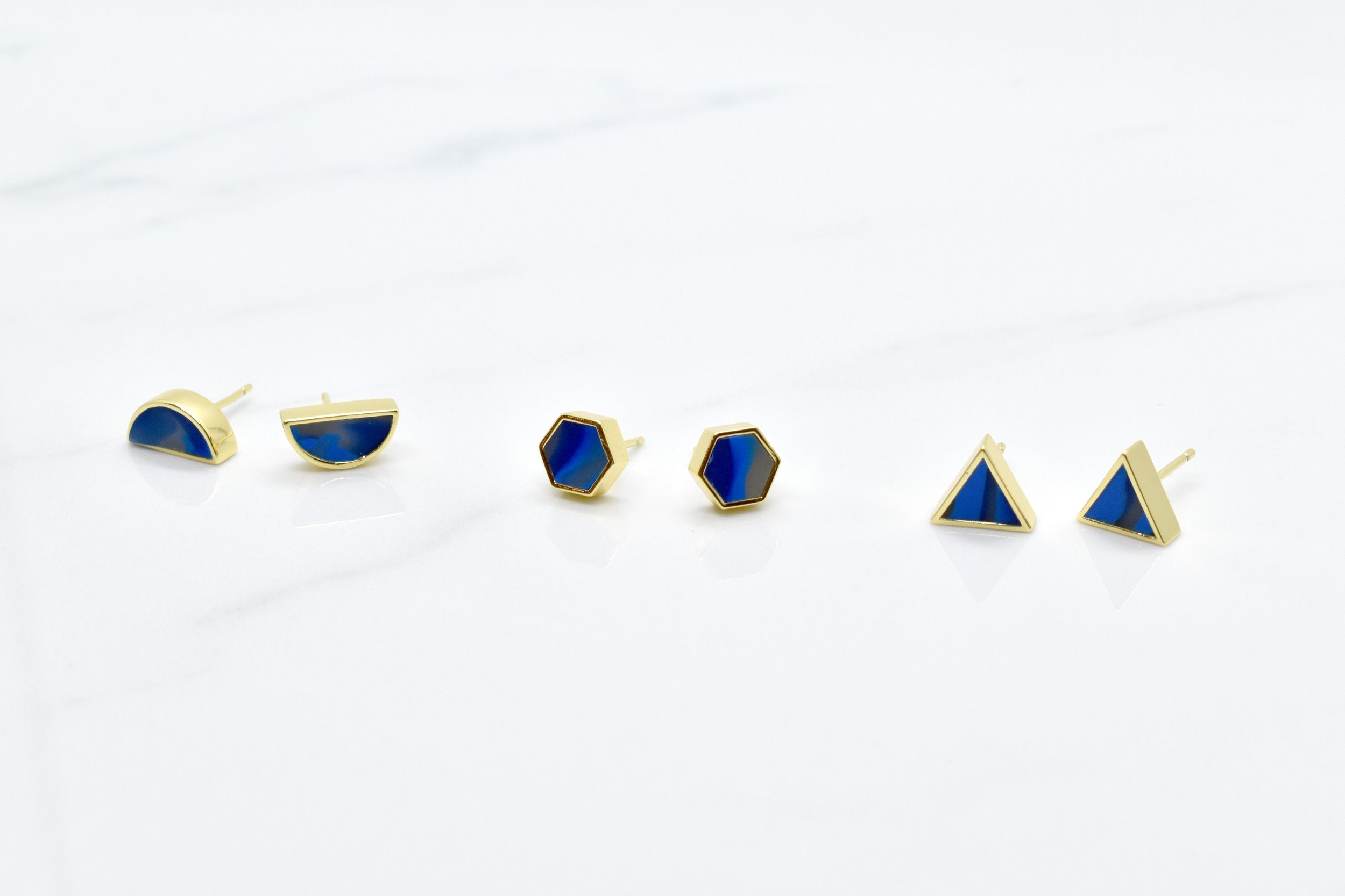 a set of three pairs of gold geometric stud earrings. One set of triangle studs, one set of gold hexagon earrings and one set of half moon stud earrings, all in sapphire gemstone that's been marble