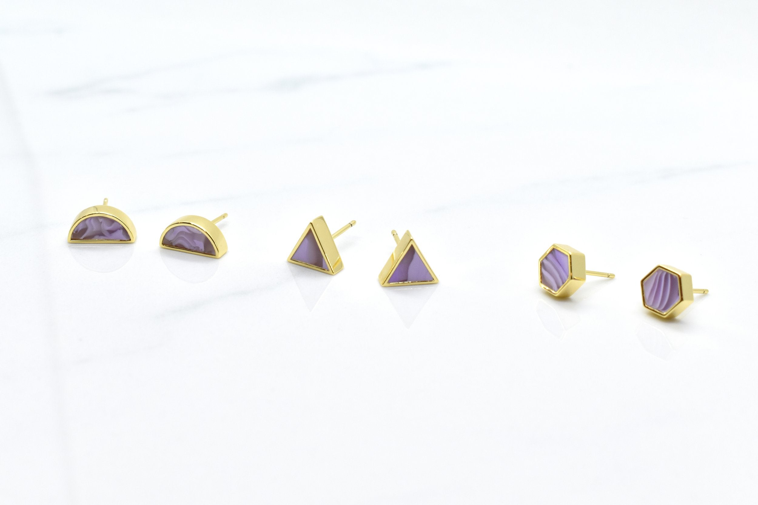 three pairs of modern and minimal pastel purple earrings in three shapes: hexagon studs, triangle studs, and half-moon studs with gold details.