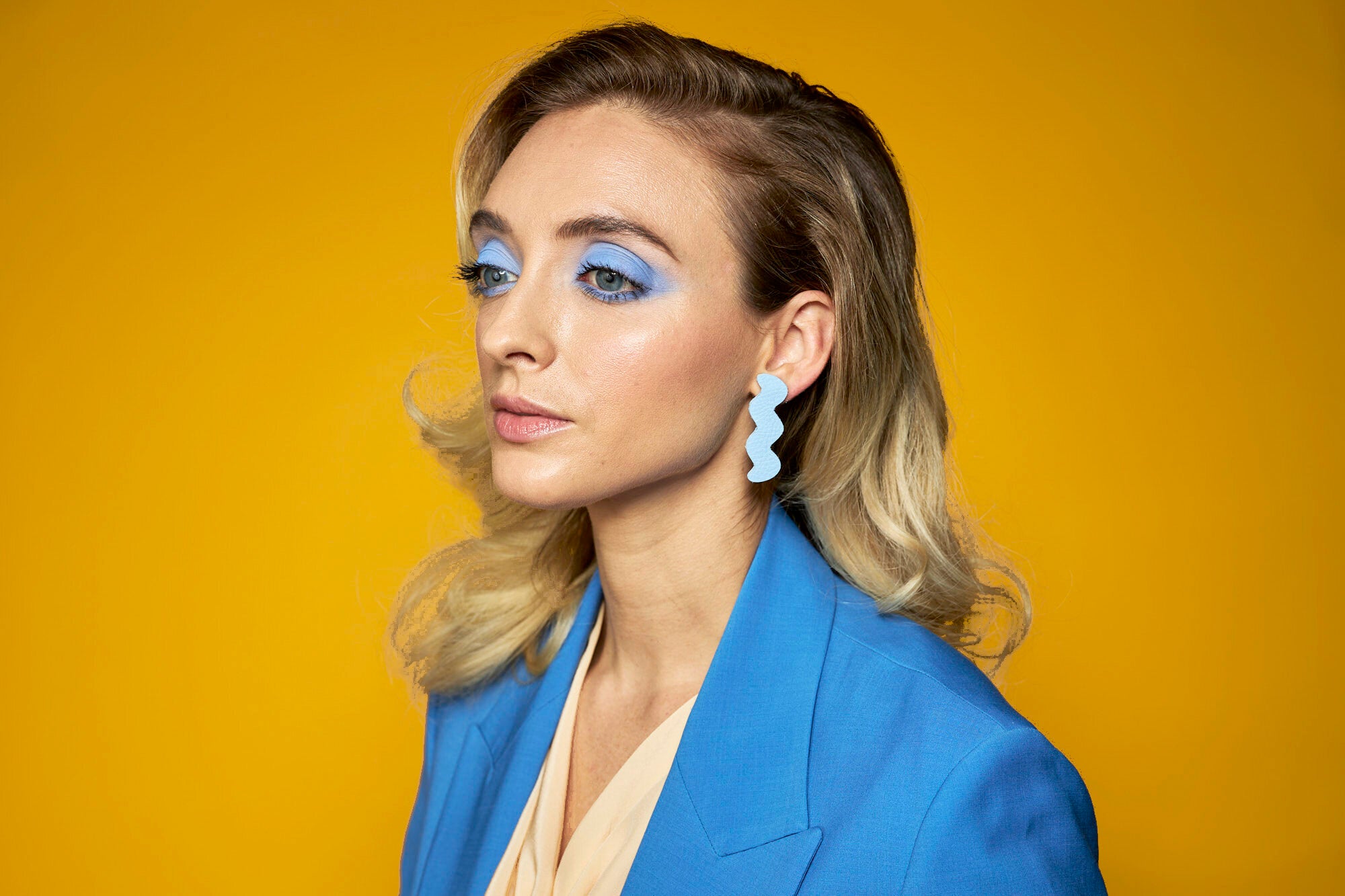 A blonde woman with bold blue makeup wears monochrome dangling periwinkle blue ear rings.