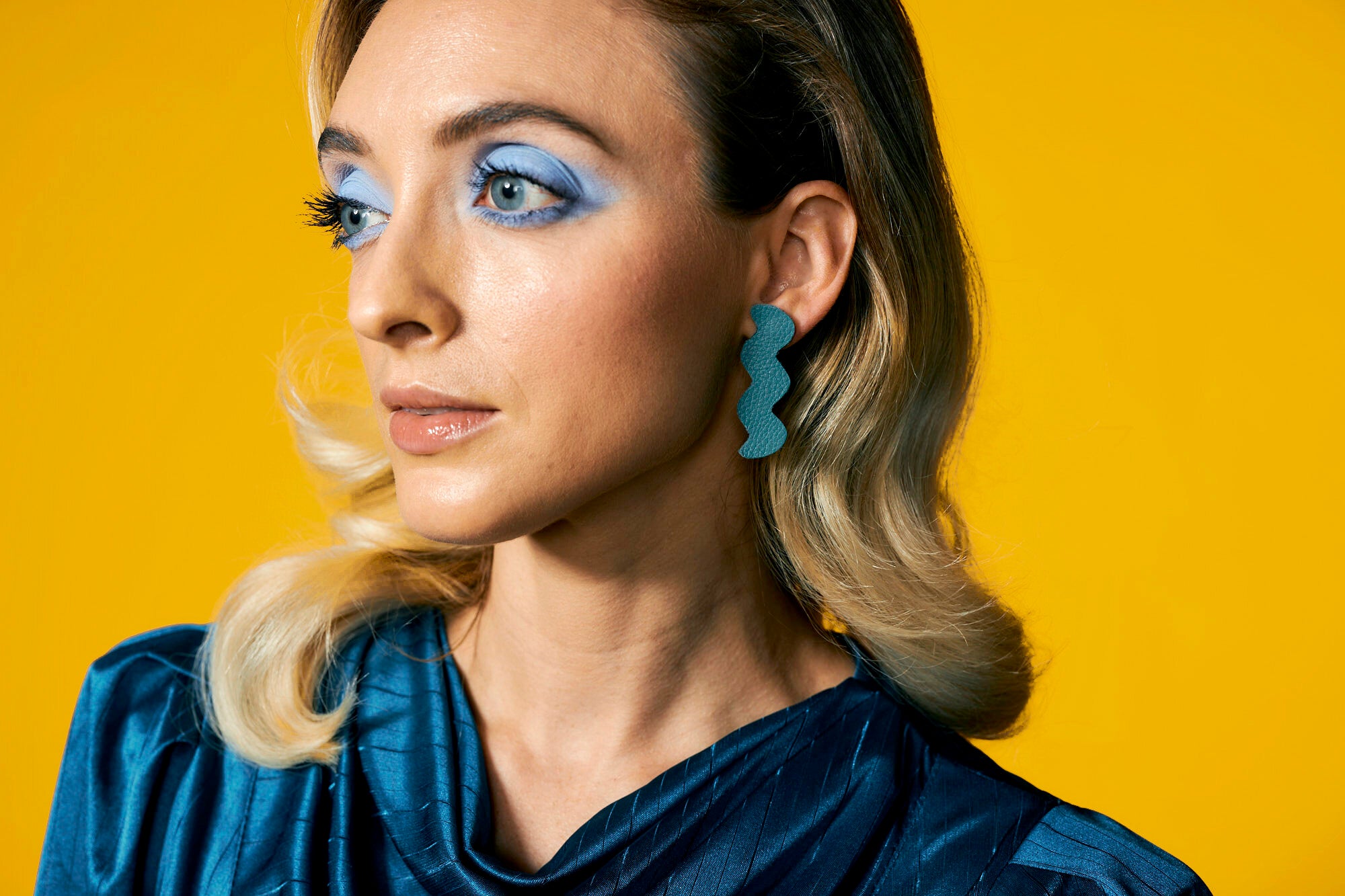 A woman dressed in turquoise styles the leather light trace earring in teal against a bold yellow background.