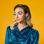 A blonde woman wears a monochromatic turquoise blue outfit wth matching teal leather statement earrings in a zig zag shape.