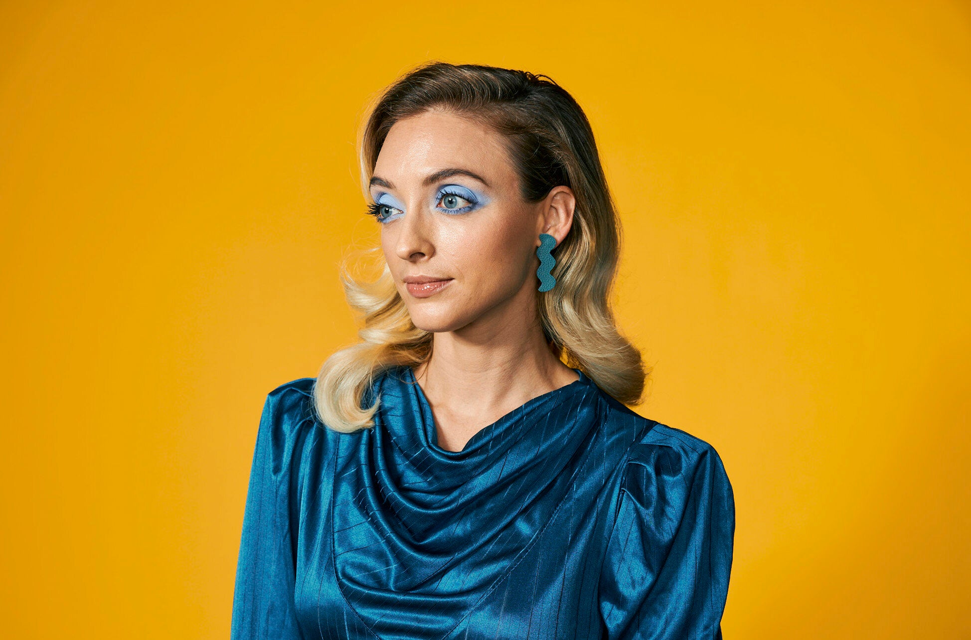 A blonde woman wears a monochromatic turquoise blue outfit wth matching teal leather statement earrings in a zig zag shape.