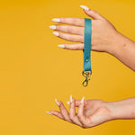 A modern key organizing wrist strap in turquoise authentic leather with gold metal detail hangs from a hand with long white fingernails.