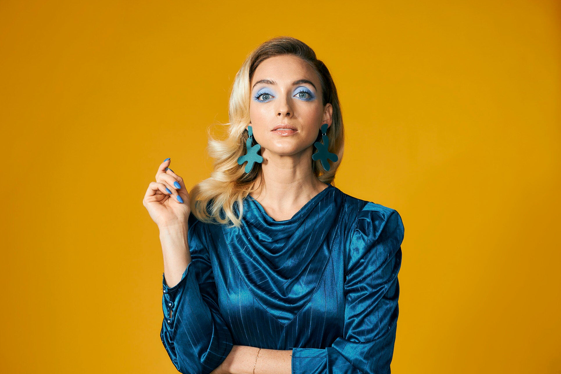 a blonde woman wears a monochromatic teal outfit with matching teal leather earrings against a yellow background.