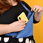 person shown putting their phone in a matisse blue sling with a gold metal zipper and crossbody strap that can also be worn as a wristlet clutch
