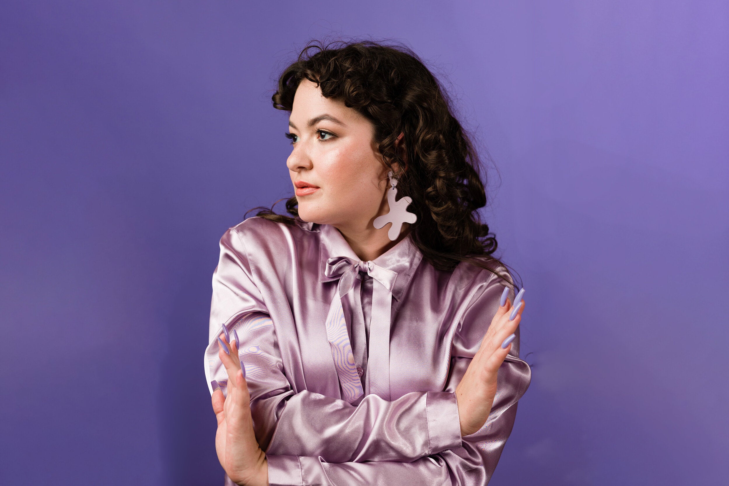 A dark haired young woman poses in front of a purple background wearing a lavender top and matching lavender leather earrings.