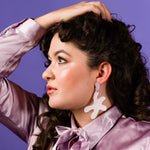 a woman with dark curly hair and dewy makeup wears a shiny lavender blouse and matching lavender leather earrings