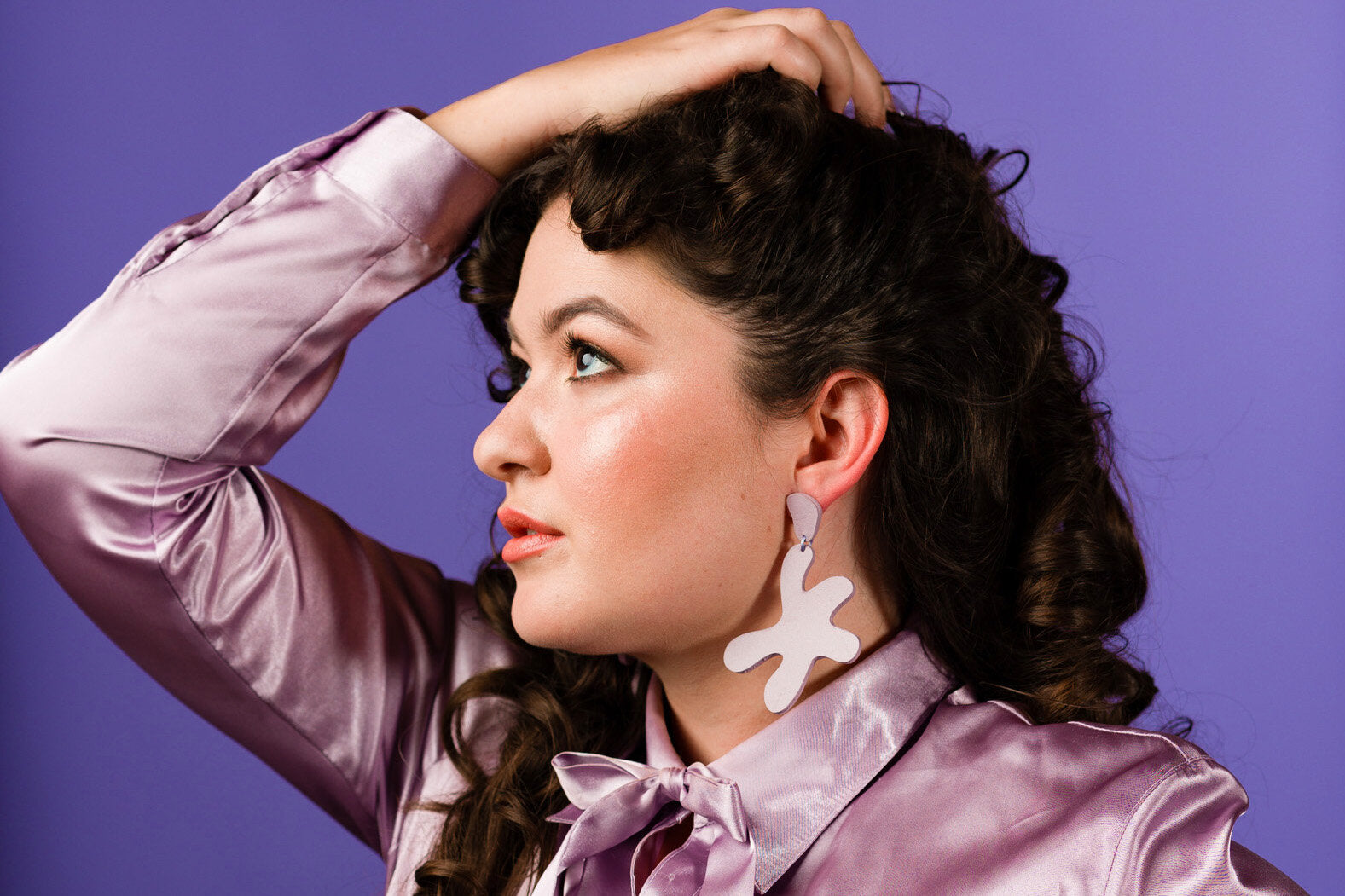 a woman with dark curly hair and dewy makeup wears a shiny lavender blouse and matching lavender leather earrings